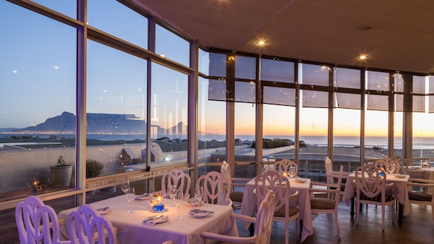Ein Restaurant mit gedeckten Tischen, großen Fenstern mit Blick auf das Meer und ferne Berge, sowie einem modernen Interieur.