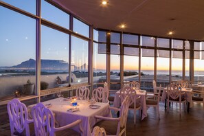 A restaurant with tables set for dining, large windows offering a view of the ocean and distant mountains, and a modern interior design. - Dolphin Beach Hotel (Cape Town)