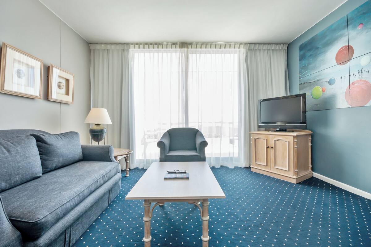 Ein Hotelzimmer mit grauem Sofa, einem kleinen Tisch, einem Fernseher auf einer Holzkommode und einem großen Fenster mit transparenten Vorhängen.