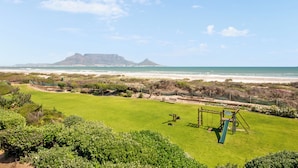 Eine Rasenfläche mit Schaukel und Rutsche, im Hintergrund ein Strand und ein Berg.