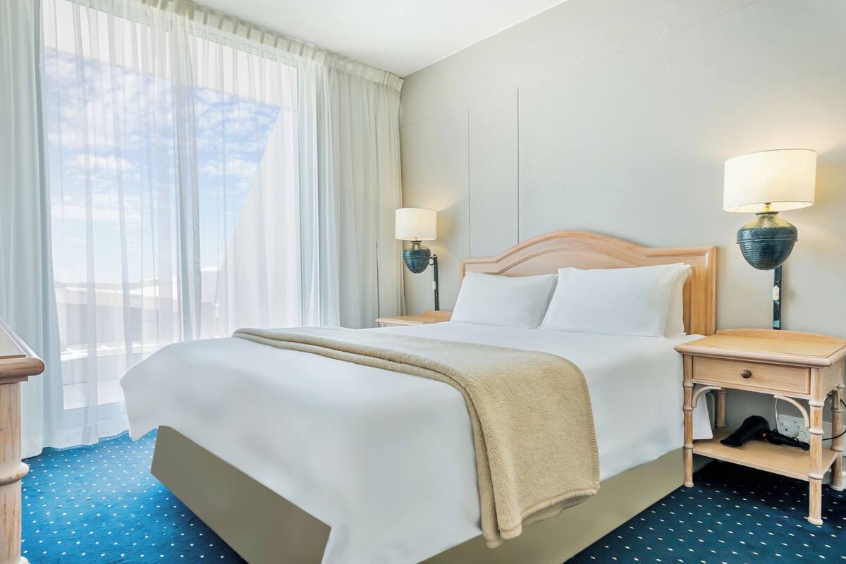 Ein Hotelzimmer mit einem Bett, zwei Nachttischen mit Lampen, einem Fenster mit Vorhängen und einem blauen Teppich mit Punkten.
