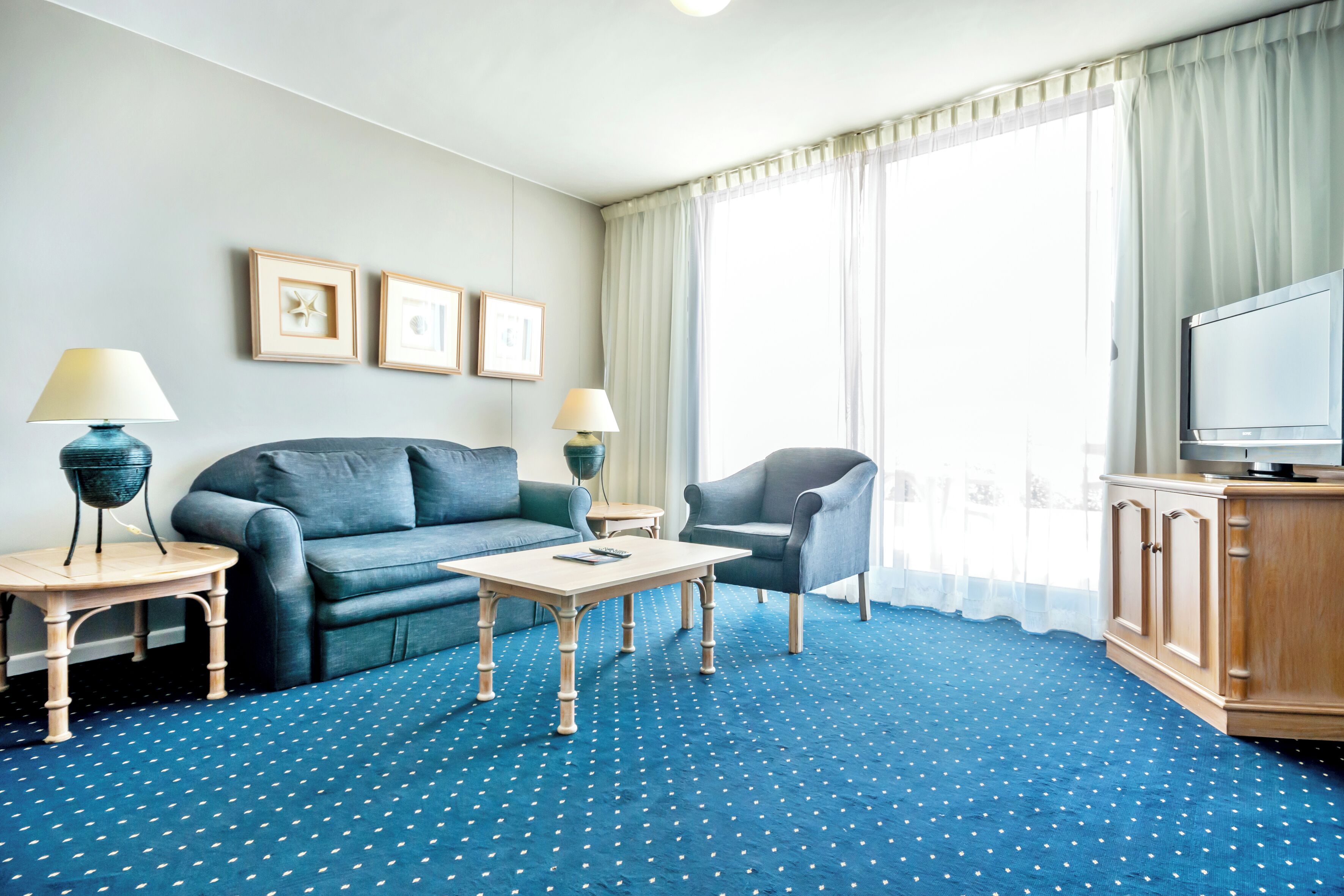 Una habitación de hotel con alfombra azul de lunares, un sofá, una silla, una mesita, dos lámparas, un televisor y cuadros en la pared.