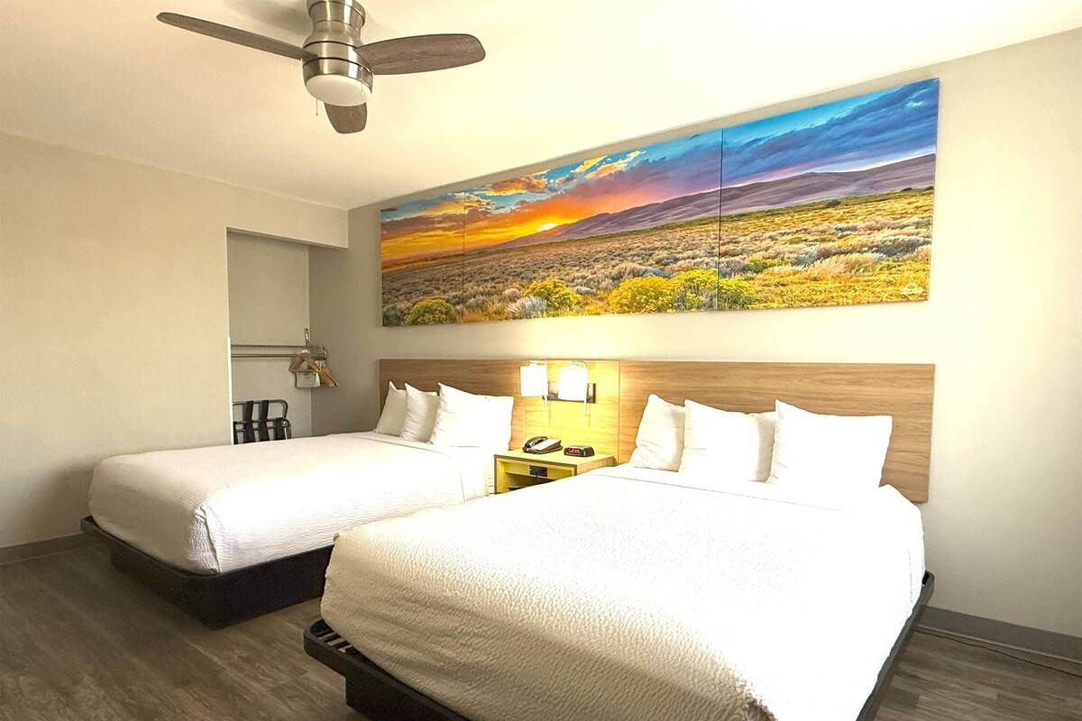 Une chambre d’hôtel avec deux lits, un ventilateur de plafond, un tableau représentant un paysage au coucher du soleil et une table de chevet avec une lampe.