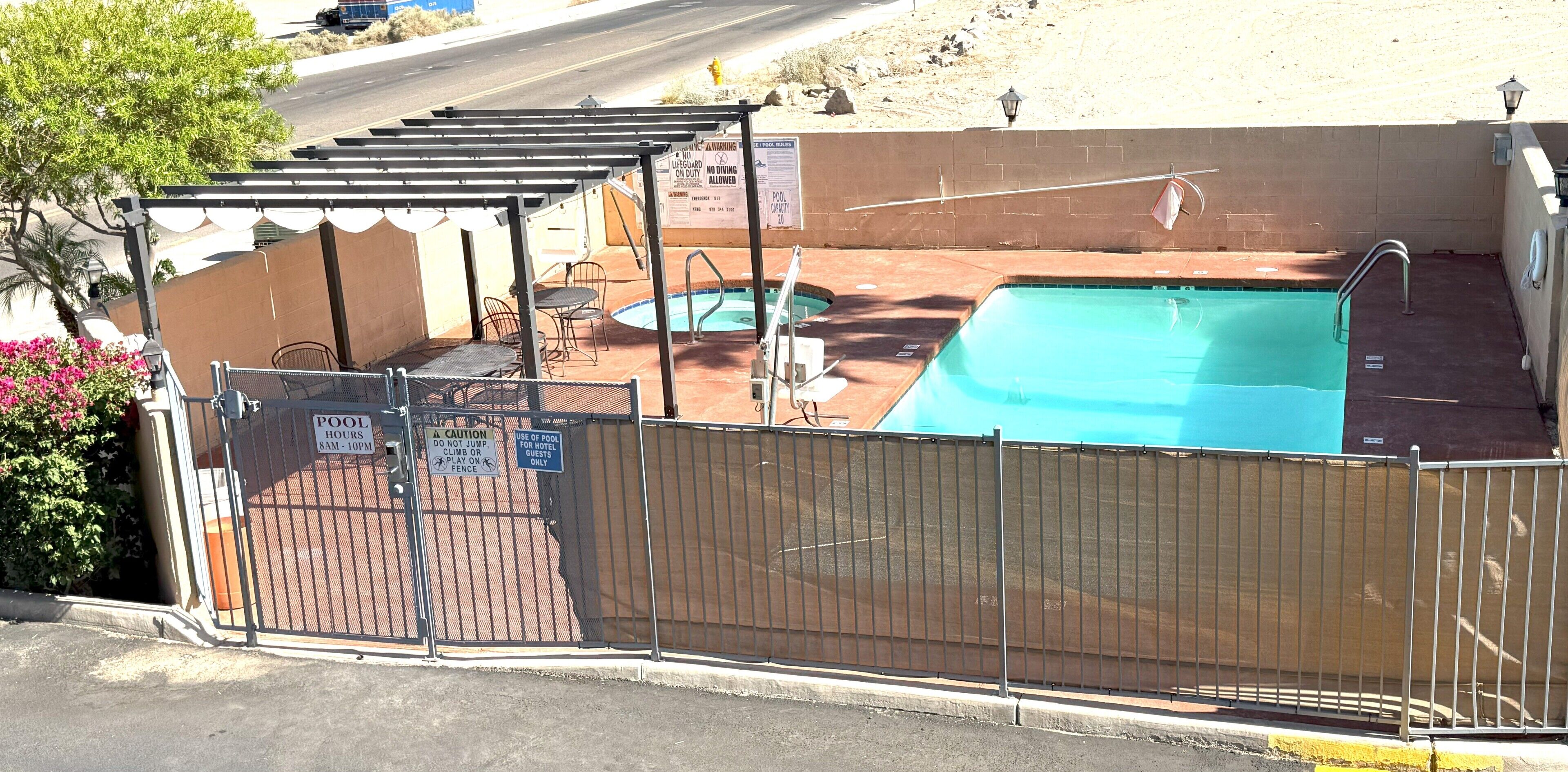 Un espace extérieur avec piscine, entouré d’une clôture, comprenant un coin salon couvert et un panneau indiquant les heures d’ouverture de la piscine.