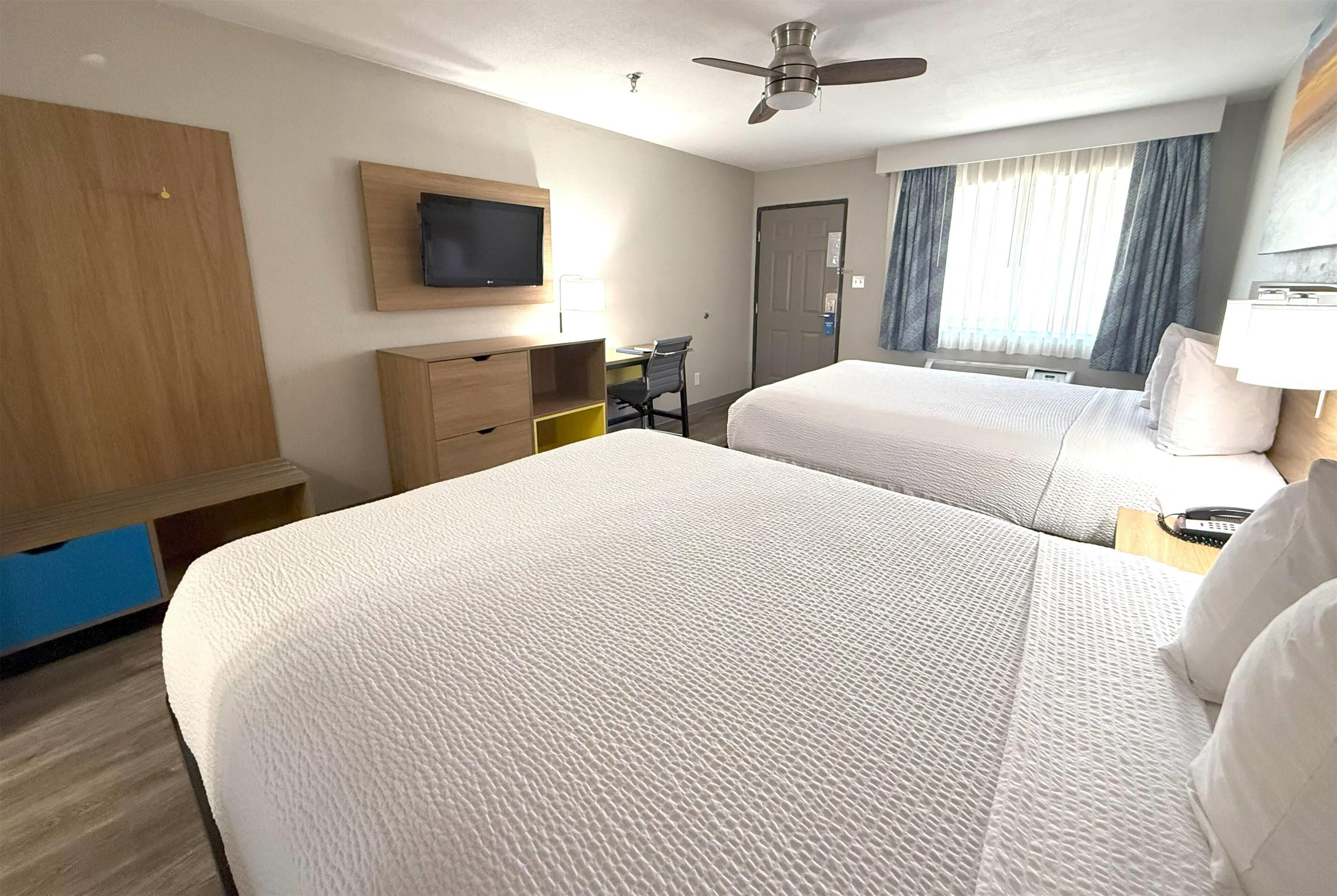 A hotel room with two beds, a desk, a chair, a television, and a ceiling fan.