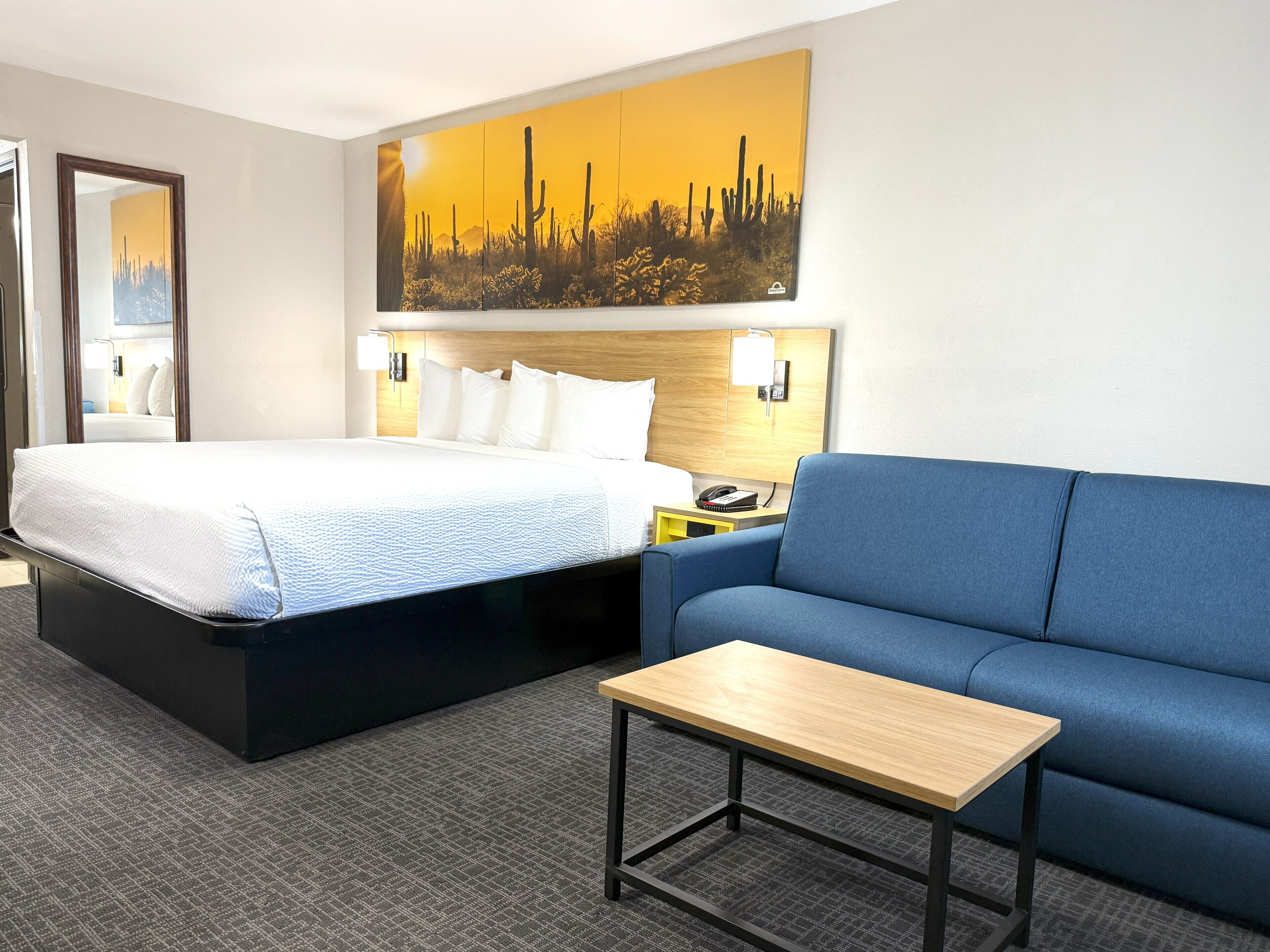 A hotel room with a bed, a blue sofa, a small table, and a painting of a desert landscape on the wall.