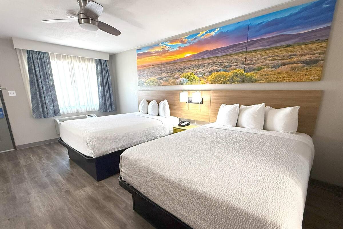 Une chambre d’hôtel avec deux lits, un ventilateur de plafond, un grand tableau représentant un paysage et une fenêtre avec des rideaux.