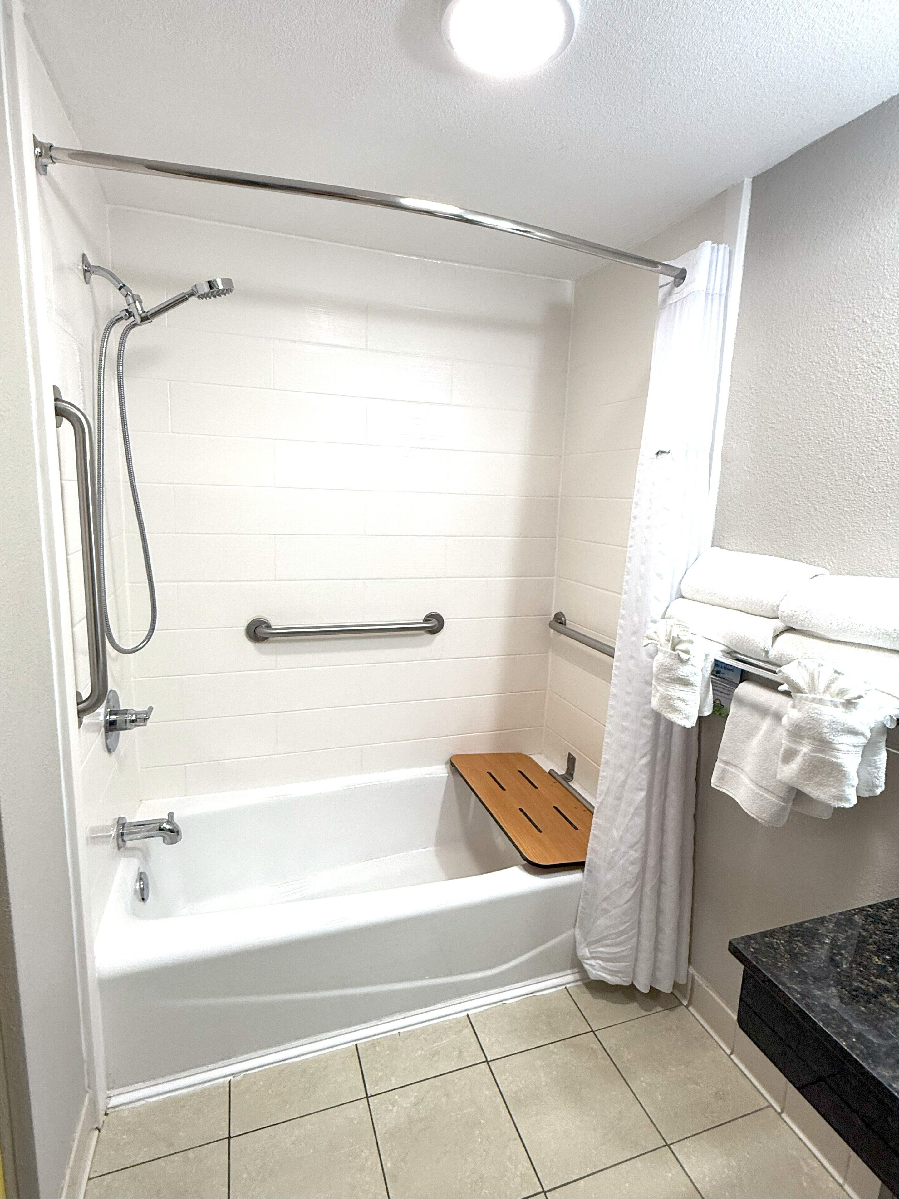 A bathroom with a white bathtub, grab bars, a wooden bath tray, a towel rack, and a black countertop.