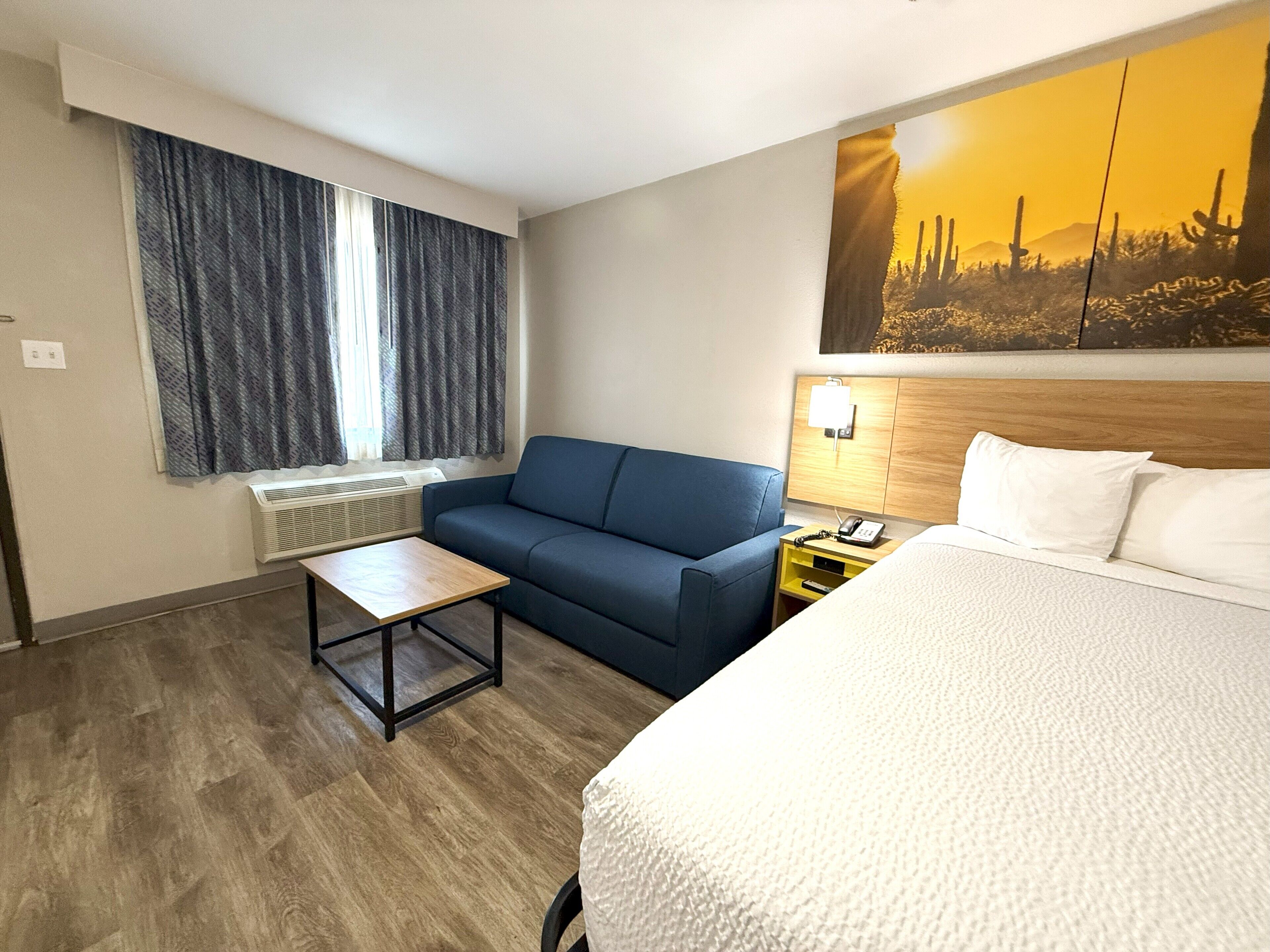 A hotel room with a bed, a sofa, a small table, a nightstand with a lamp, and a painting on the wall.