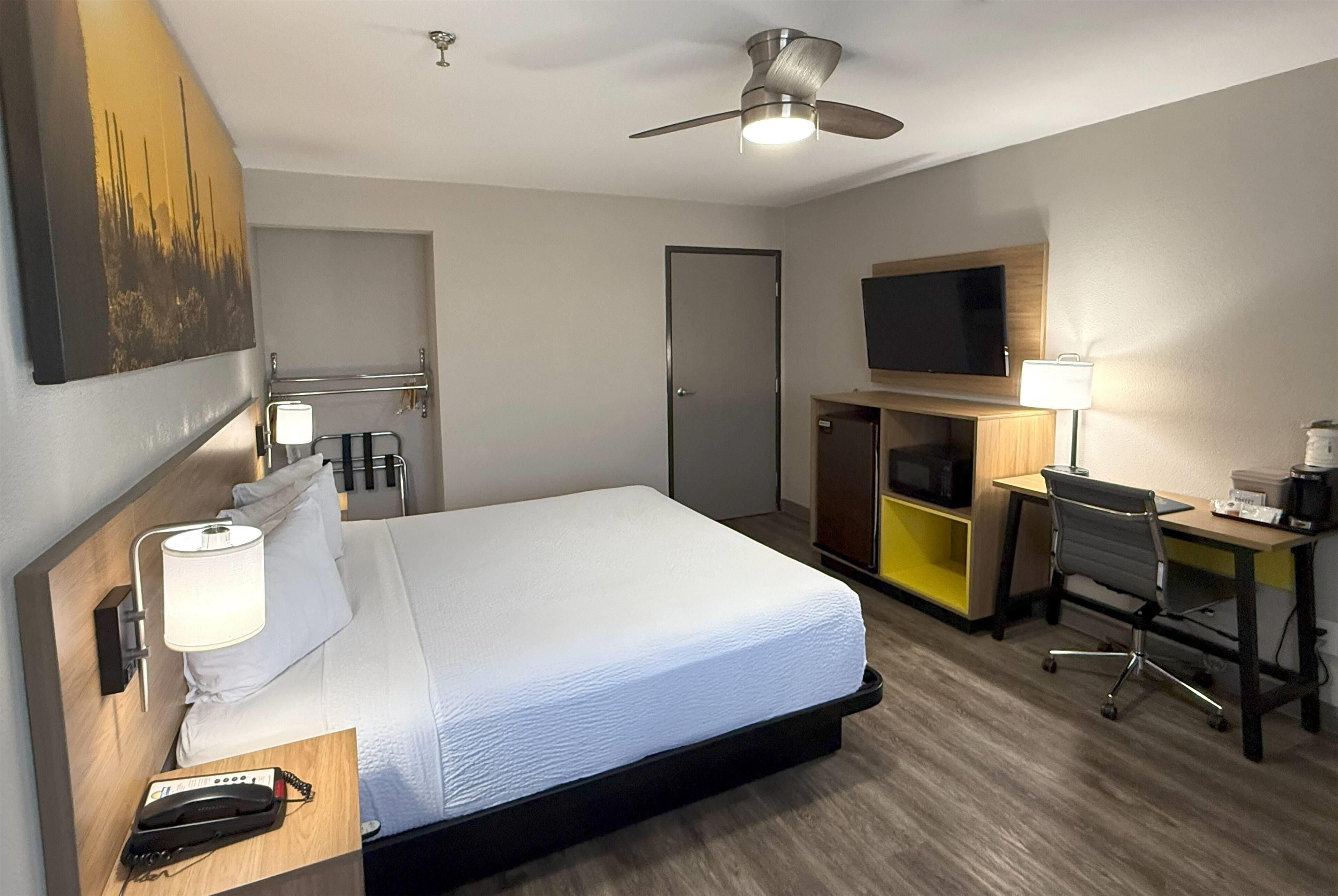 A hotel room with a bed, desk, chair, TV, ceiling fan, and a painting on the wall.