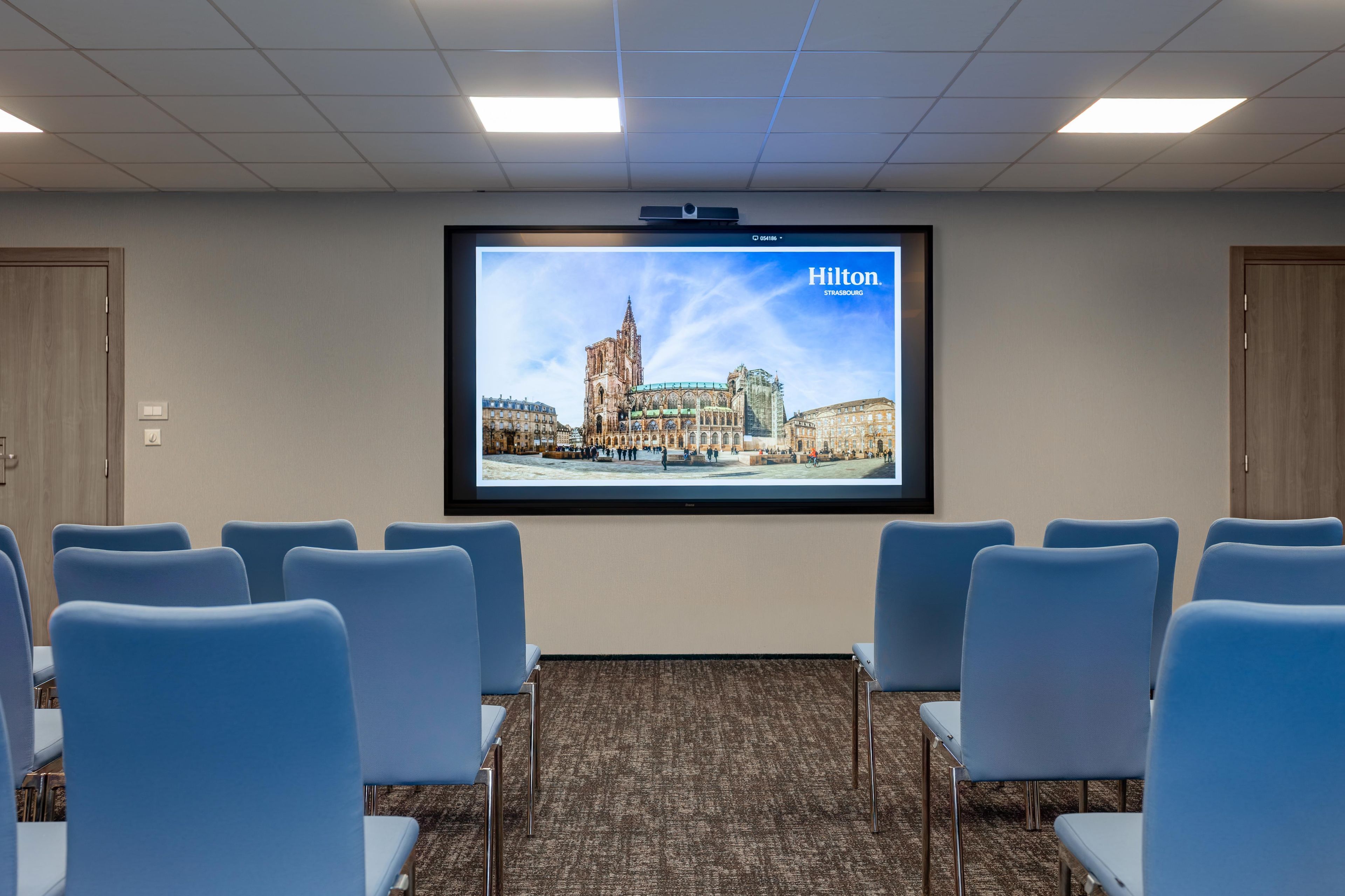 A conference room with a large screen displaying a cityscape, blue chairs arranged in rows, and a carpeted floor.