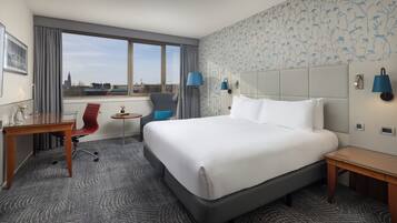 Et hotelværelse med en stor seng, et skrivebord, en stol, et lille bord og udsigt til bybilledet gennem vinduet.