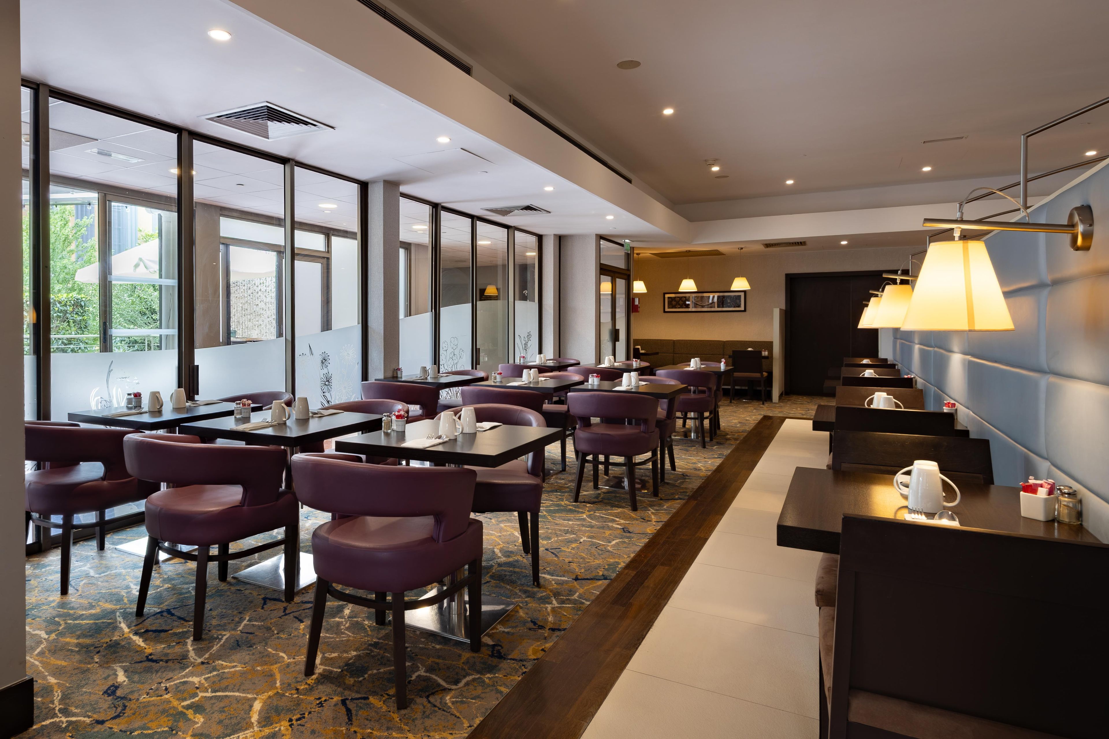 Une salle à manger d’hôtel avec des tables et des chaises, de grandes fenêtres et un tapis à motifs.