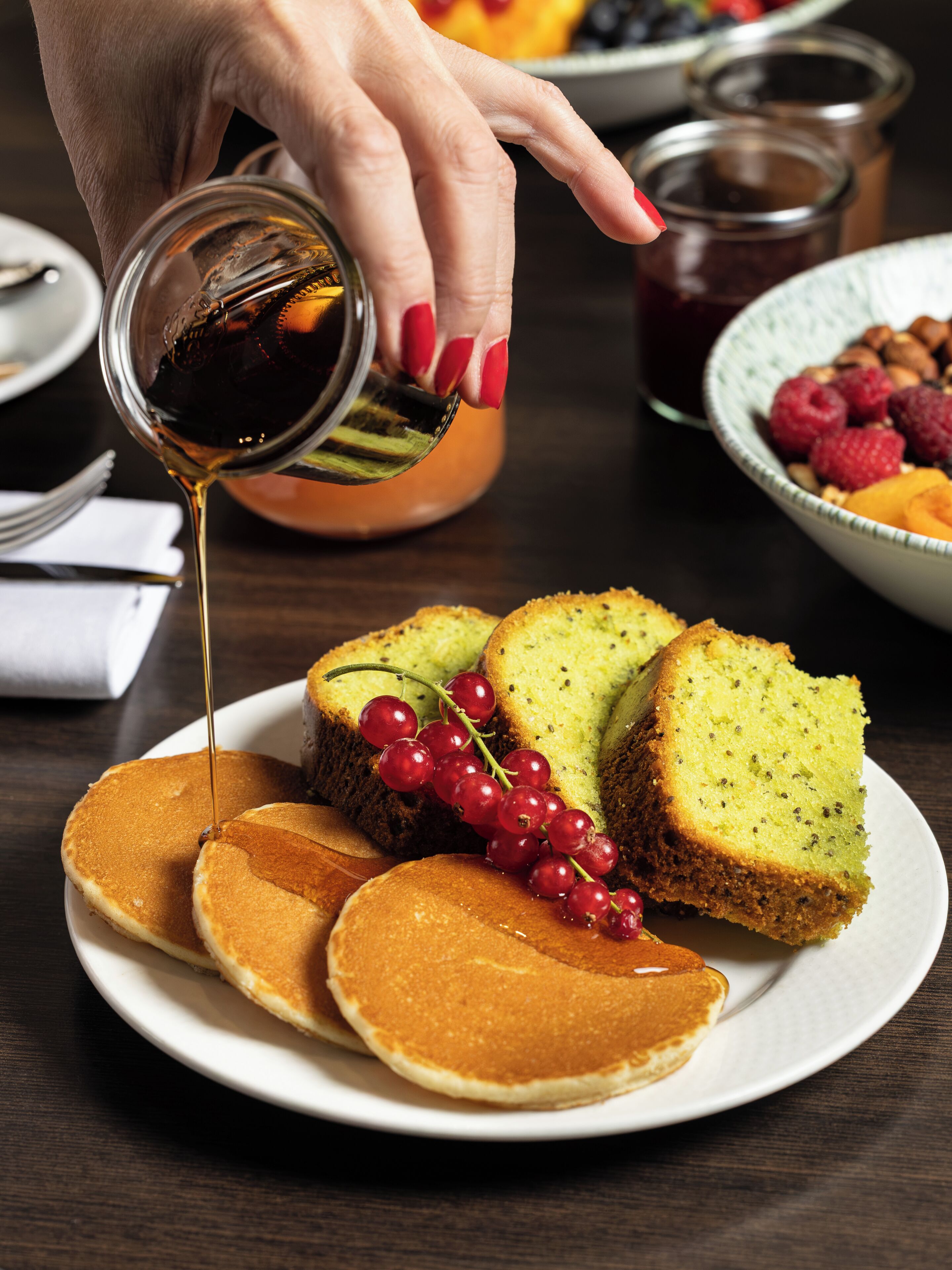 A plate with pancakes, cake, and red currants, with syrup being poured over them.