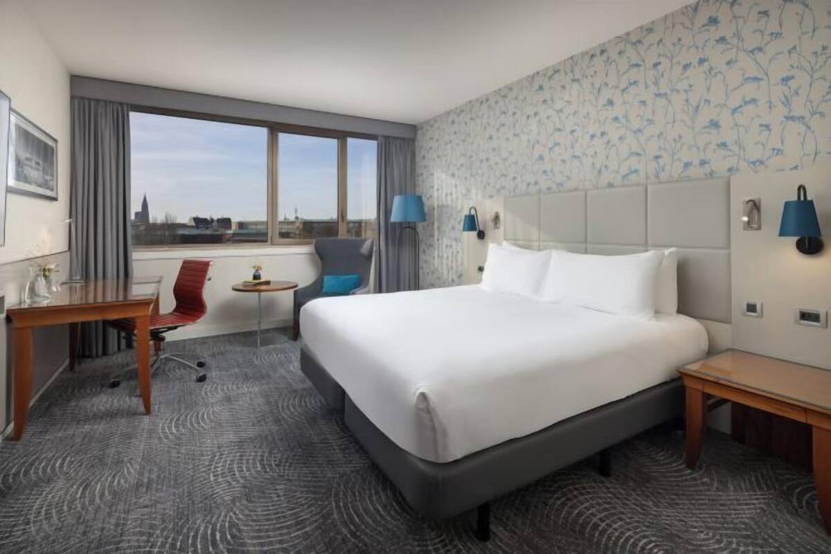 A hotel room with a large bed, a desk, a chair, a small table, and a view of the city through large windows.