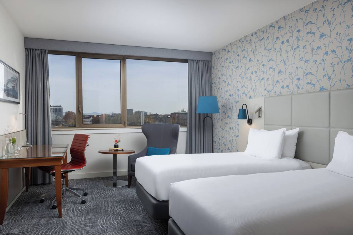 A hotel room with two beds, a desk, a chair, a small table, and a view of the city.