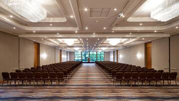 Un salón de conferencias con filas de sillas, una alfombra estampada, candelabros y un gran ventanal al frente.