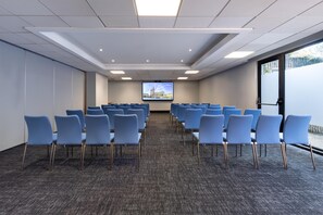 Un salón de conferencias con filas de sillas azules, piso alfombrado y una pantalla grande al frente.