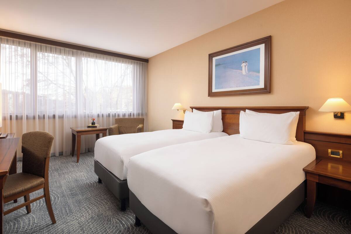 A hotel room with two beds, a desk, a chair, and a window with sheer curtains.