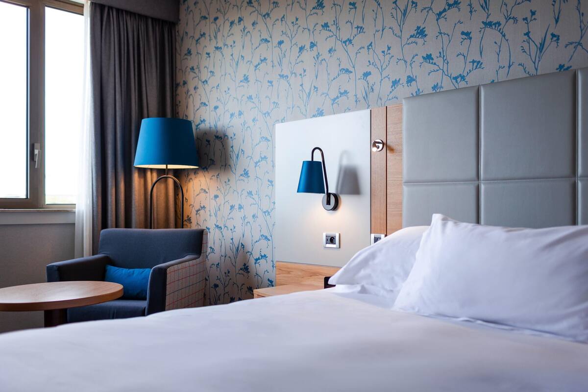 A hotel room with a bed, a chair, a small table, a lamp, and a wall-mounted light fixture.