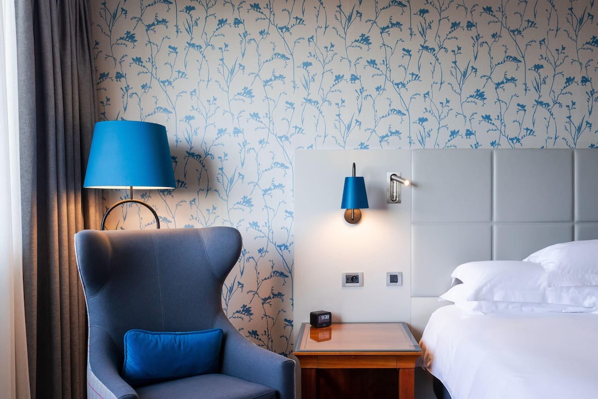 A hotel room with a blue armchair, a bedside table with a lamp, a bed with white linens, and floral wallpaper.