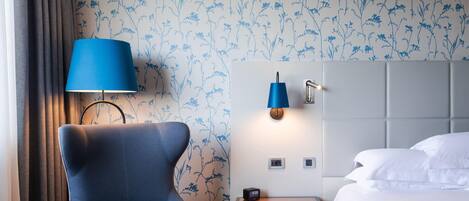 A hotel room with a blue armchair, a bedside table with a lamp, a bed with white linens, and floral wallpaper.