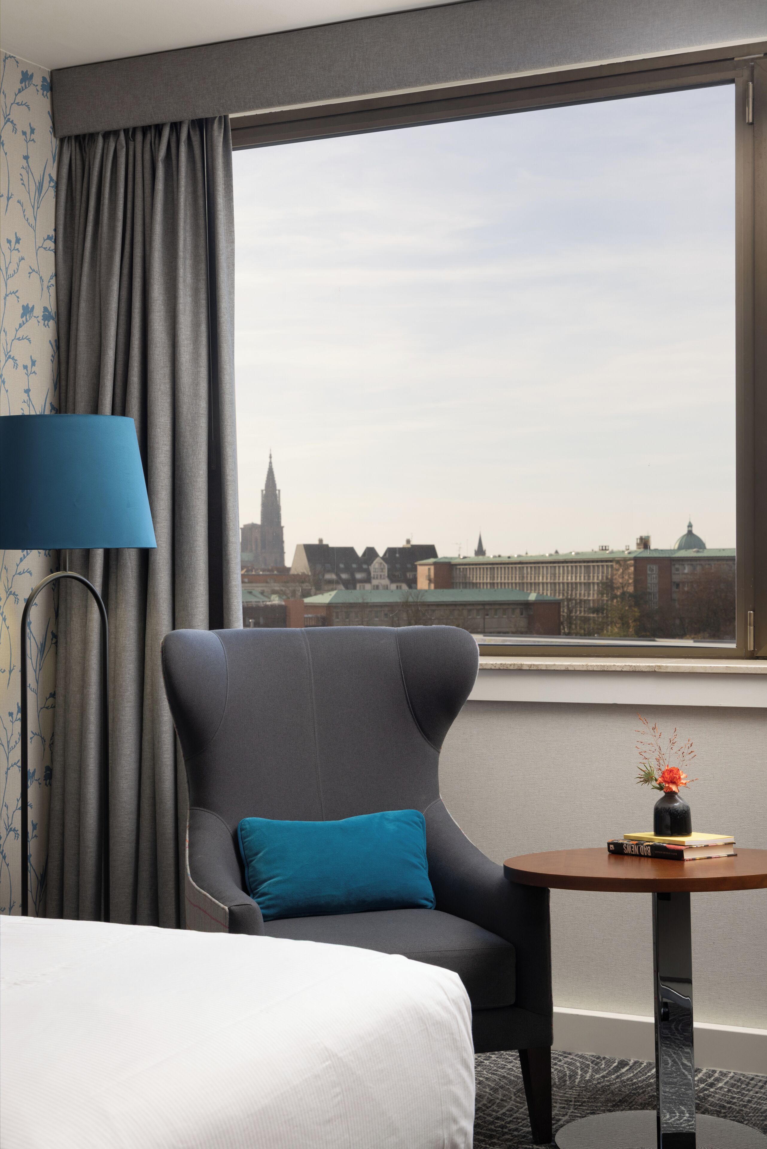 Habitación de hotel con una cama, un sillón gris, una mesita redonda con un jarrón y vistas a un paisaje urbano a través de un ventanal amplio.