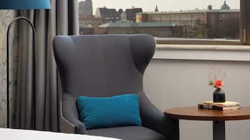 Habitación de hotel con una cama, un sillón gris, una mesita redonda con un jarrón y vistas a un paisaje urbano a través de un ventanal amplio.