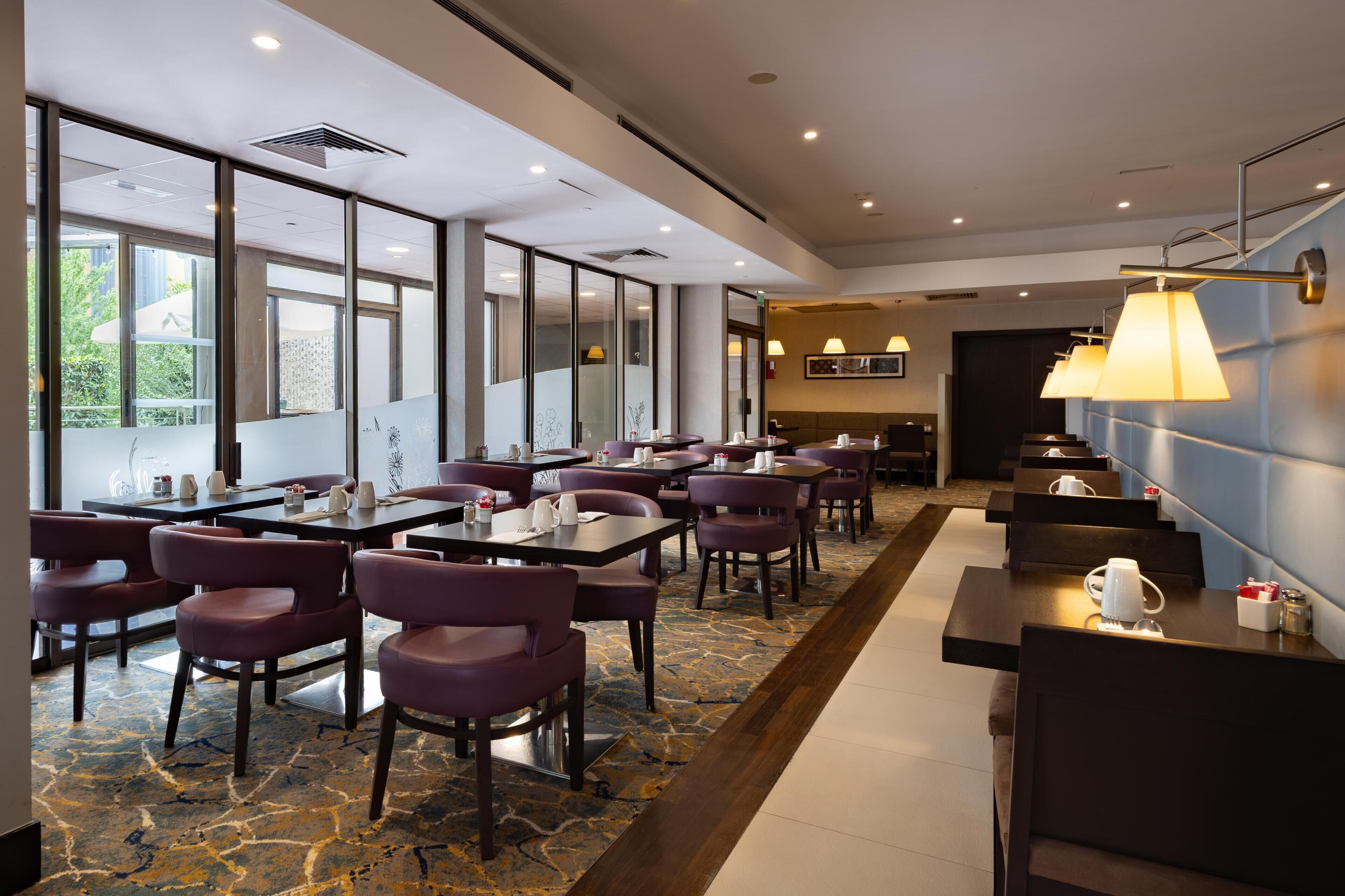 A hotel dining area with tables and chairs, large windows, and a patterned carpet.
