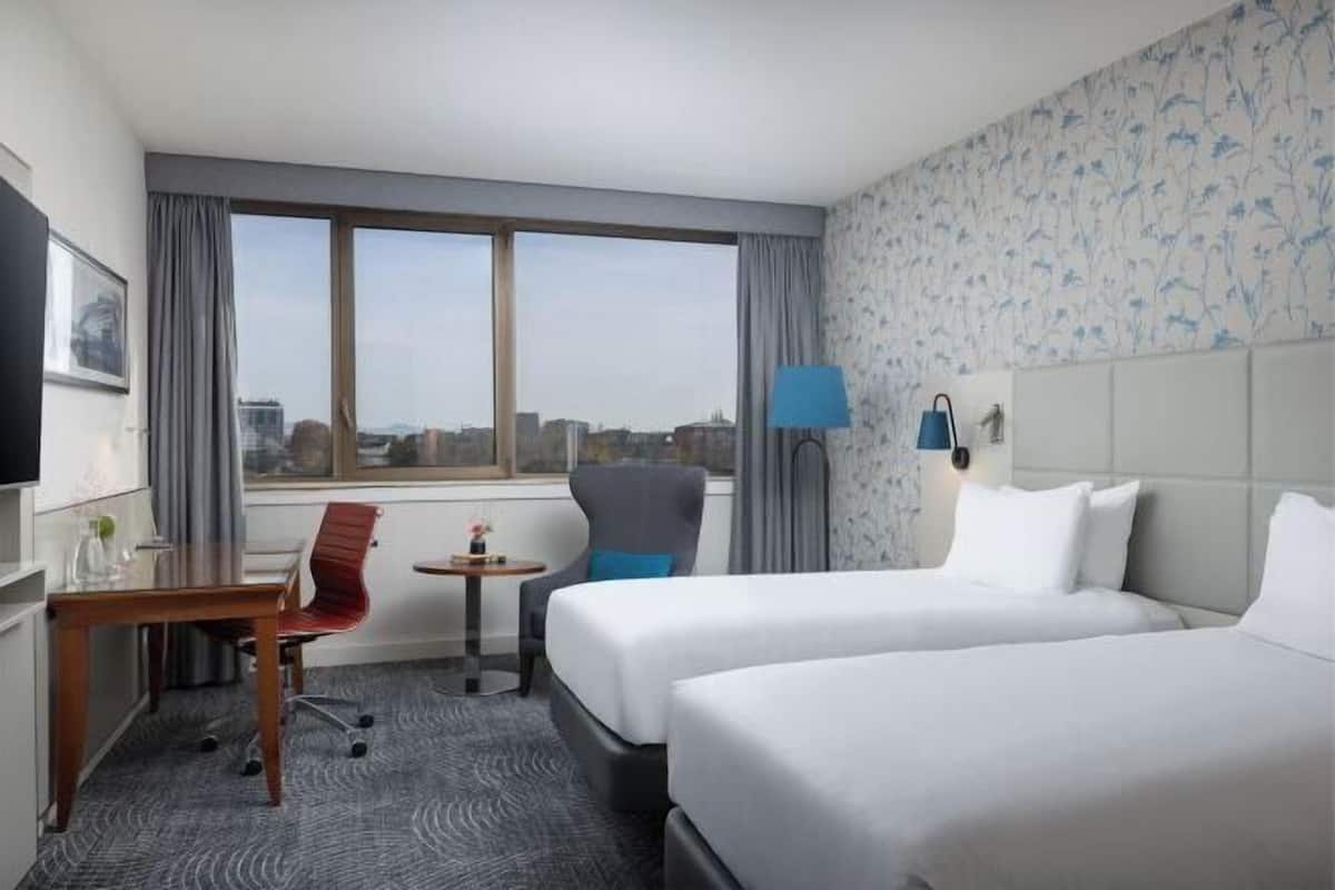 A hotel room with two beds, a desk, a chair, a lamp, and a view of the city.