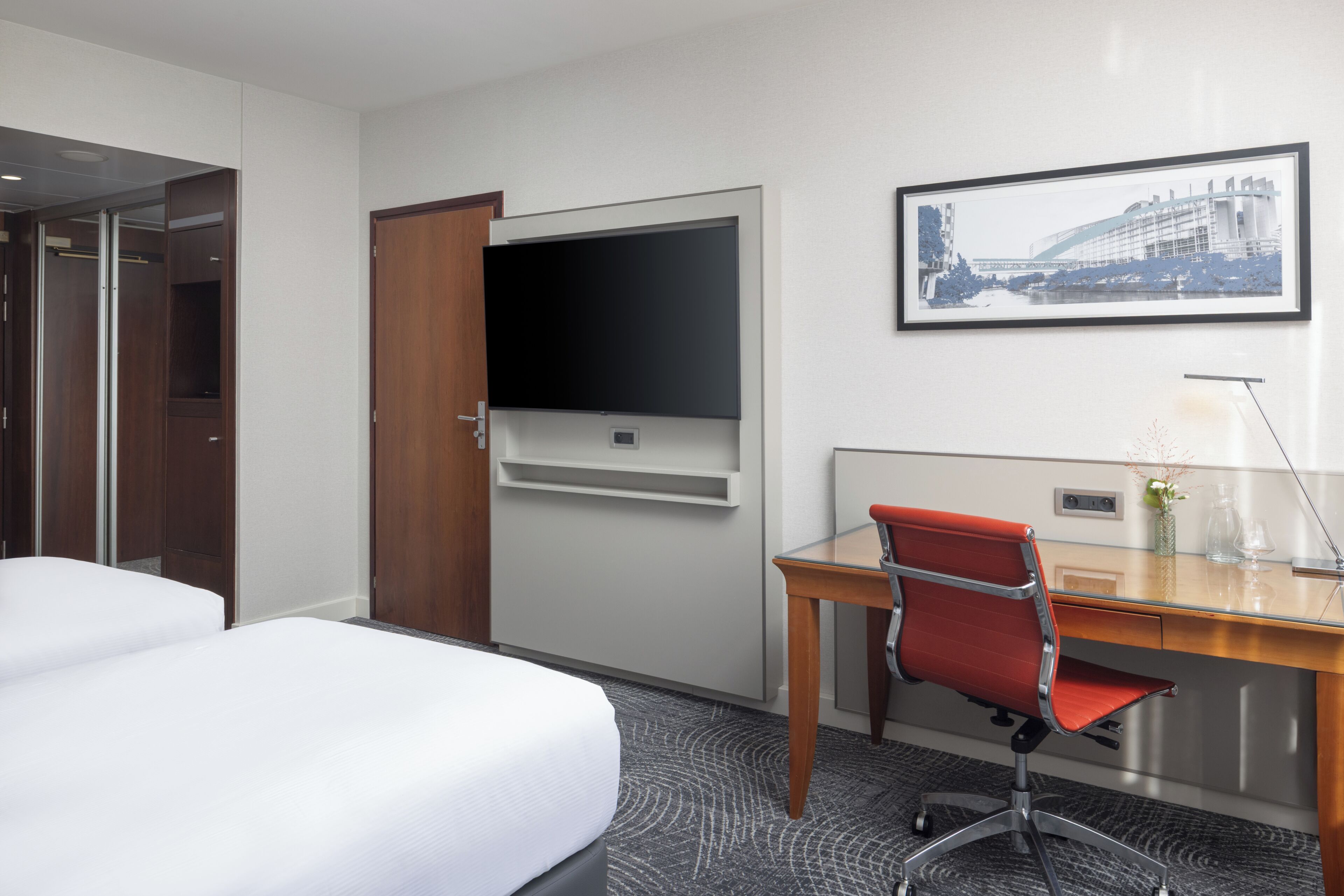 Une chambre d’hôtel avec un lit, un bureau, une chaise, une télévision et un tableau encadré au mur.