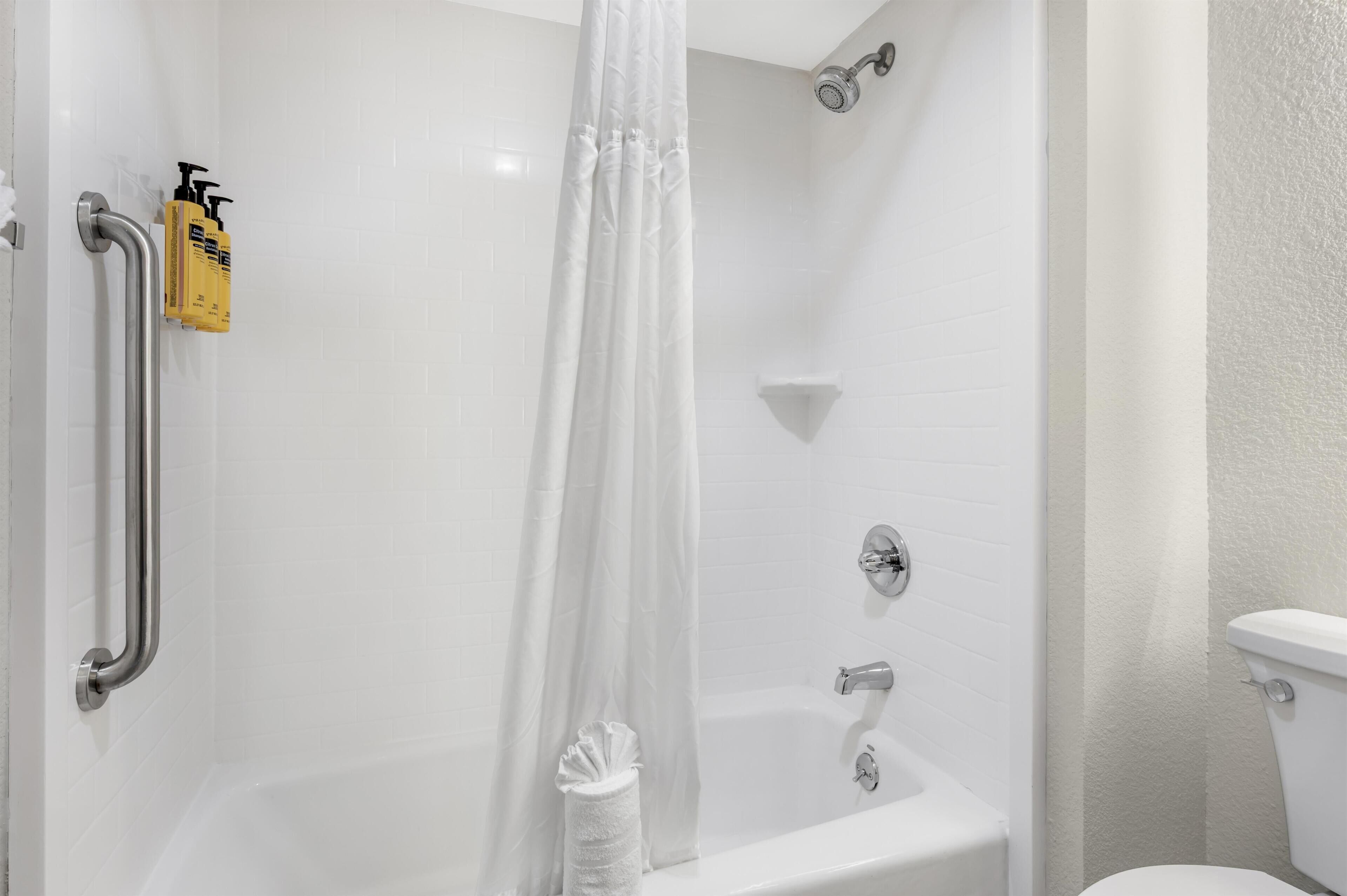 Une salle de bain avec une baignoire blanche, une douche avec un rideau blanc, des toilettes et un distributeur de gel hydroalcoolique fixé au mur.