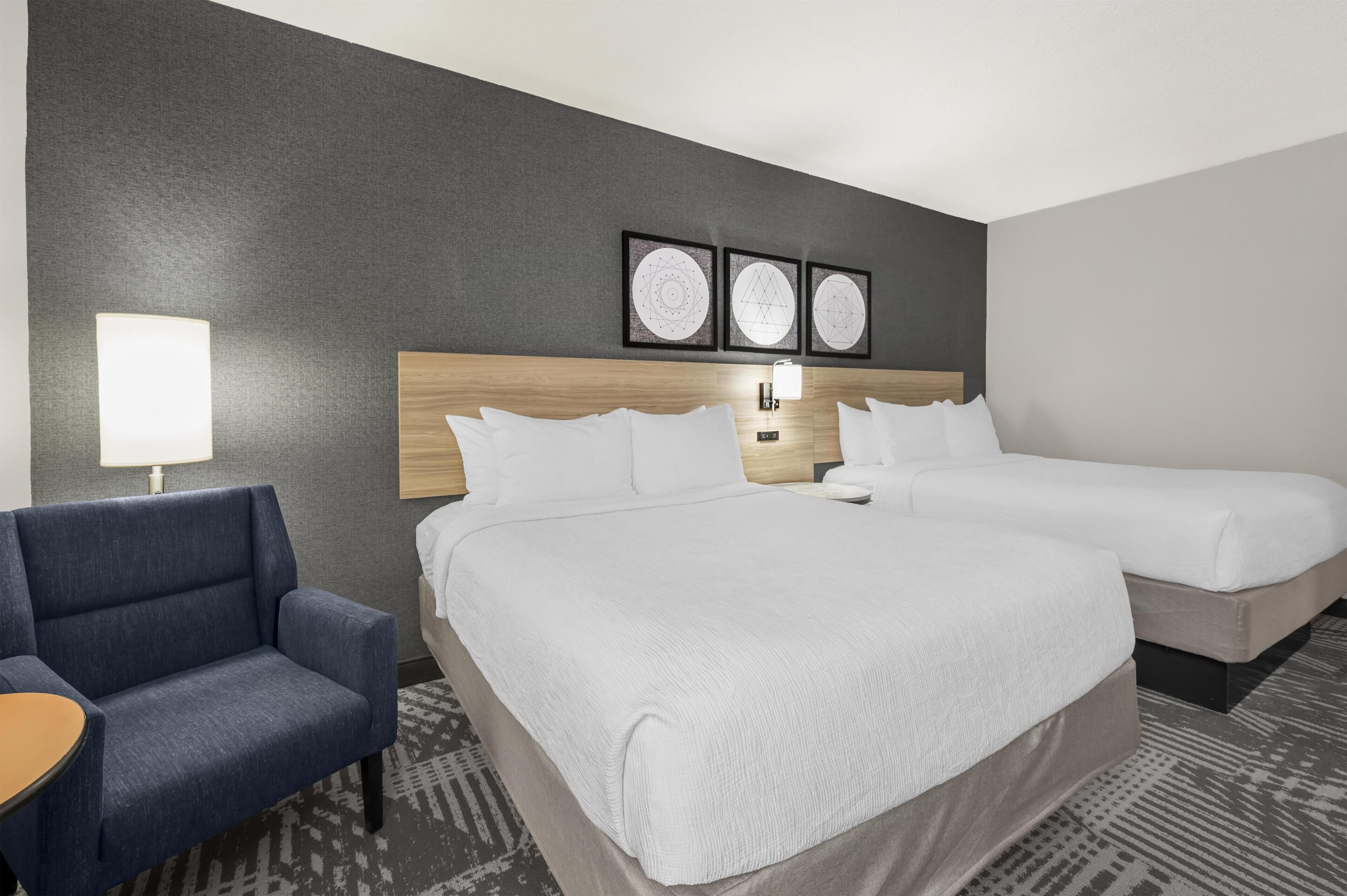 A hotel room with two beds, a desk, a chair, and a wall with three framed pictures.