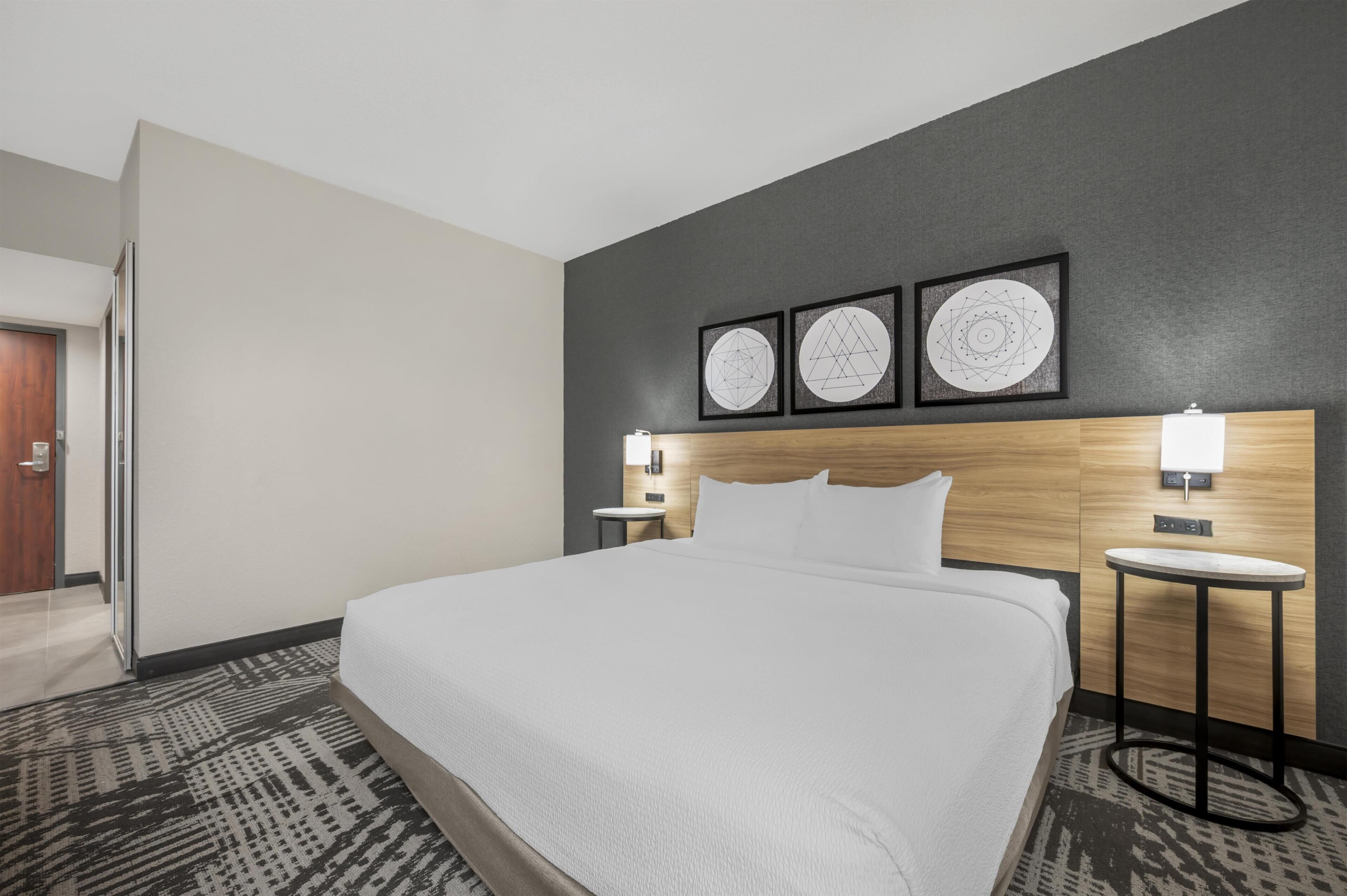 A hotel room with a large bed, a wooden headboard, a small bedside table with a lamp, and three framed wall art pieces.