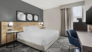 A hotel room with a bed, a desk, a chair, a TV, and framed artwork on the wall.