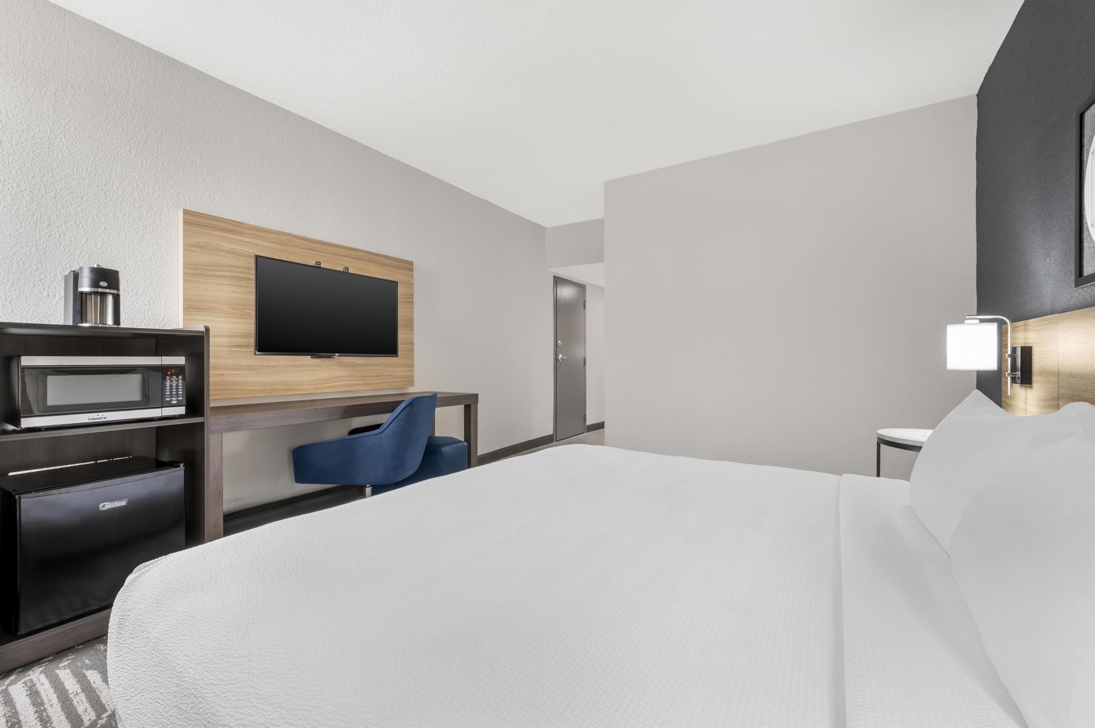 Habitación de hotel con cama, escritorio con computadora, microondas, cafetera, televisor y mesita de noche con lámpara.