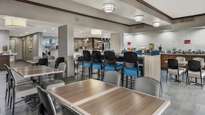 Un'area ristorazione moderna in un hotel, con diversi tavoli e sedie, un bancone della cucina e vista sulla città attraverso le finestre.