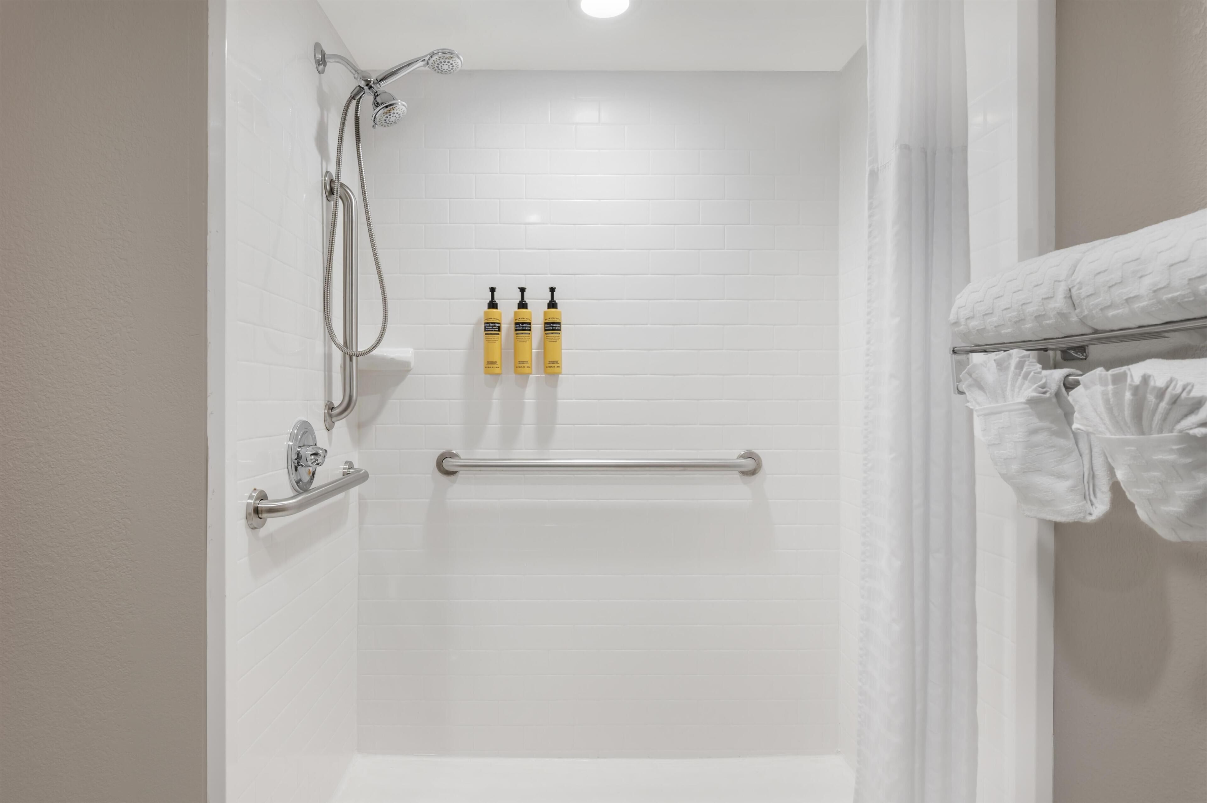 A bathroom with white tiled walls, a shower with a handheld showerhead, a grab bar, and two bottles of shampoo.