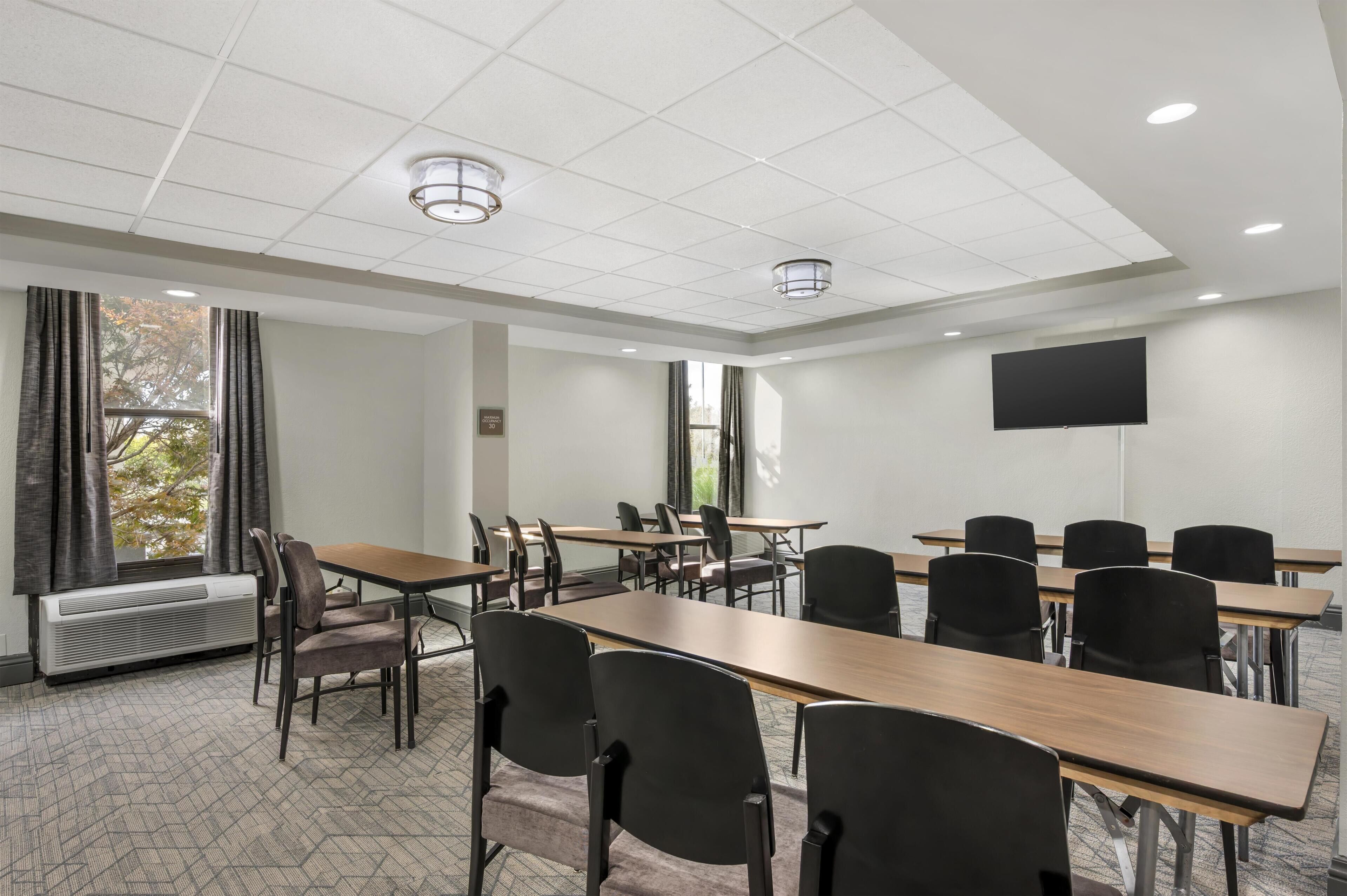 A conference room with multiple tables and chairs, a large flat-screen TV on the wall, and a window with curtains.