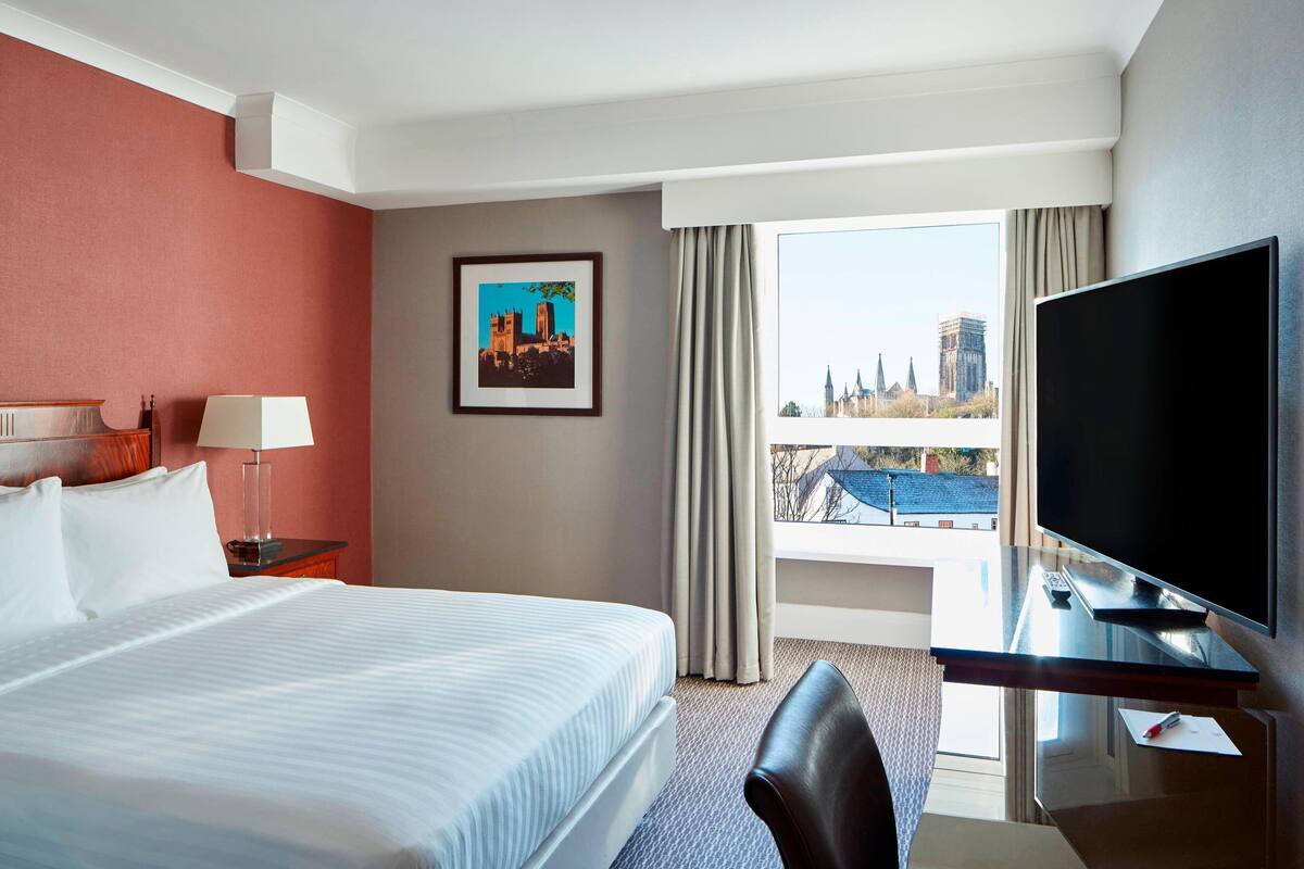 Habitación de hotel con cama, escritorio con televisor, silla, lámpara y ventana con vista a la ciudad.