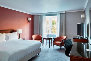 Habitación de hotel con una cama grande, dos sillones, una mesita, un televisor y una ventana con cortinas.