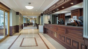 Zona de recepción de un hotel con piso de mármol, mostrador de madera y un ventanal grande.