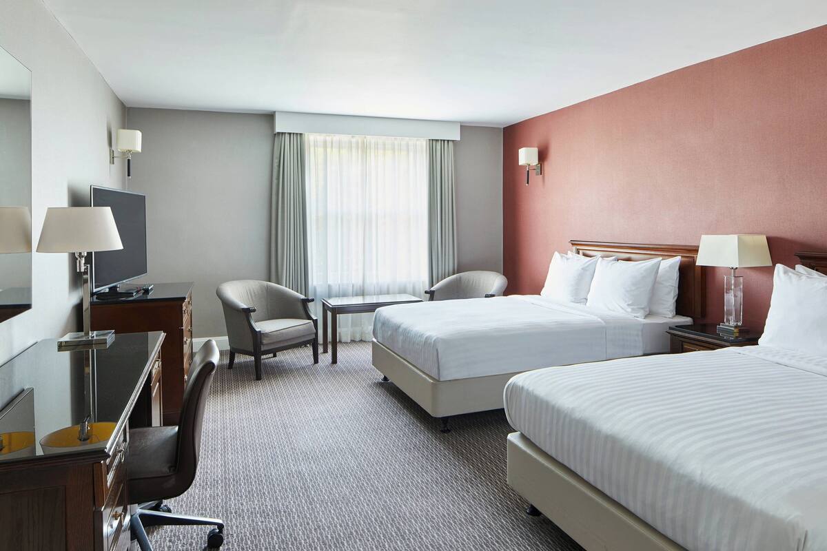 Habitación de hotel con dos camas, un escritorio con silla, televisor y ventana con cortinas.