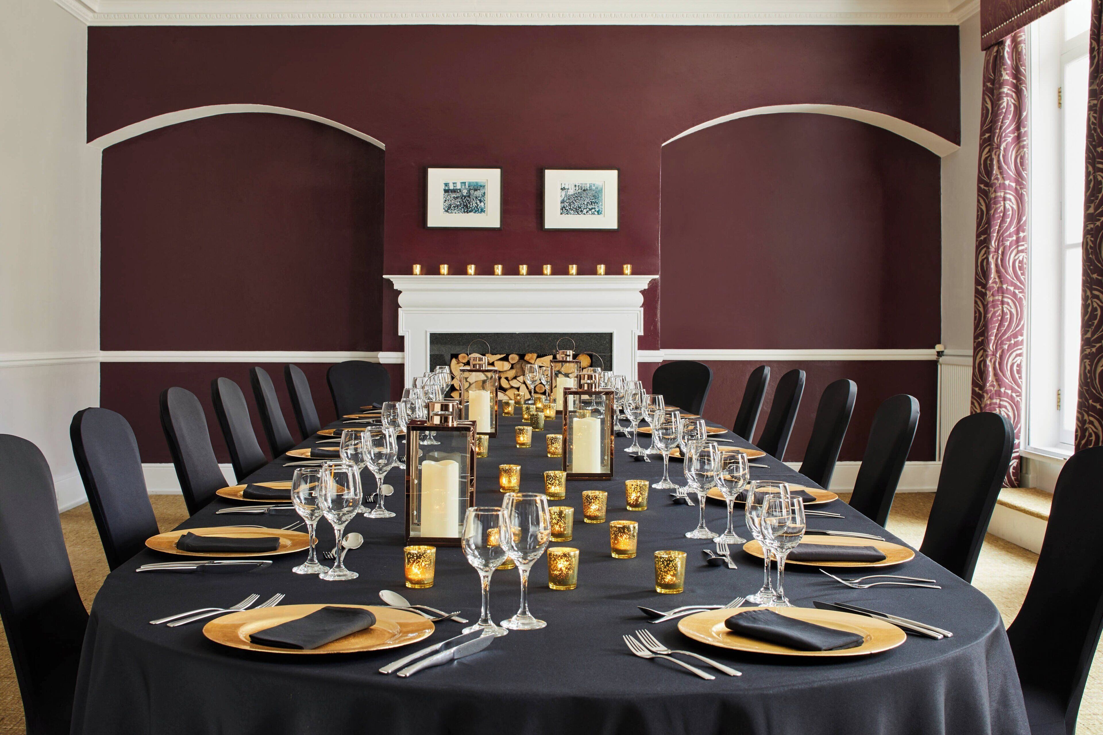 Uma sala de jantar formal com uma mesa comprida posta para refeição, com cadeiras pretas em ambos os lados, uma lareira branca e quadros decorativos nas paredes.