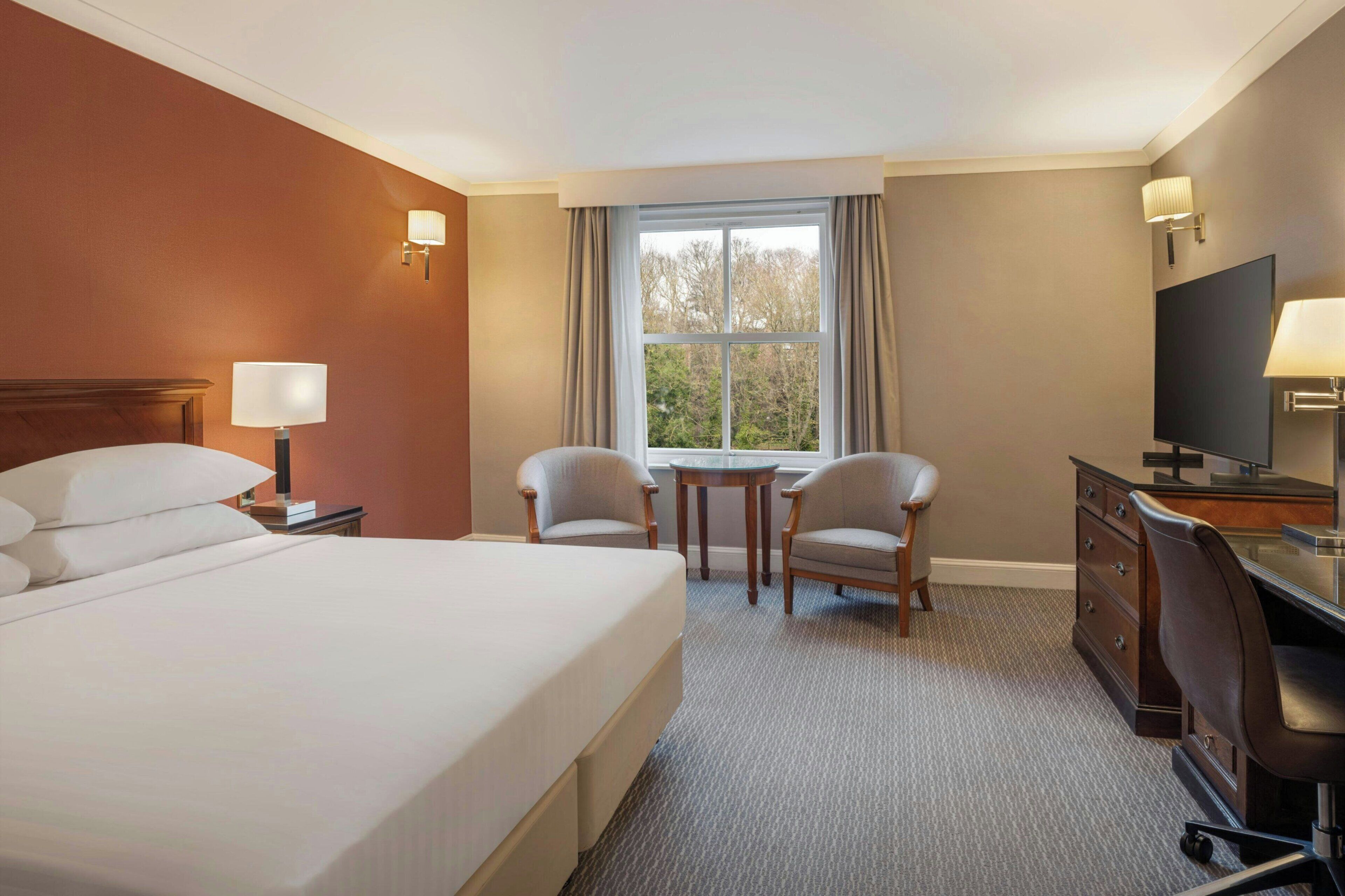 A hotel room with a large bed, a desk, two chairs, a television, and a window with curtains.
