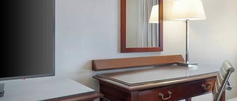 A hotel room with a wooden desk, a chair, a flat-screen TV, a mirror, and a lamp.