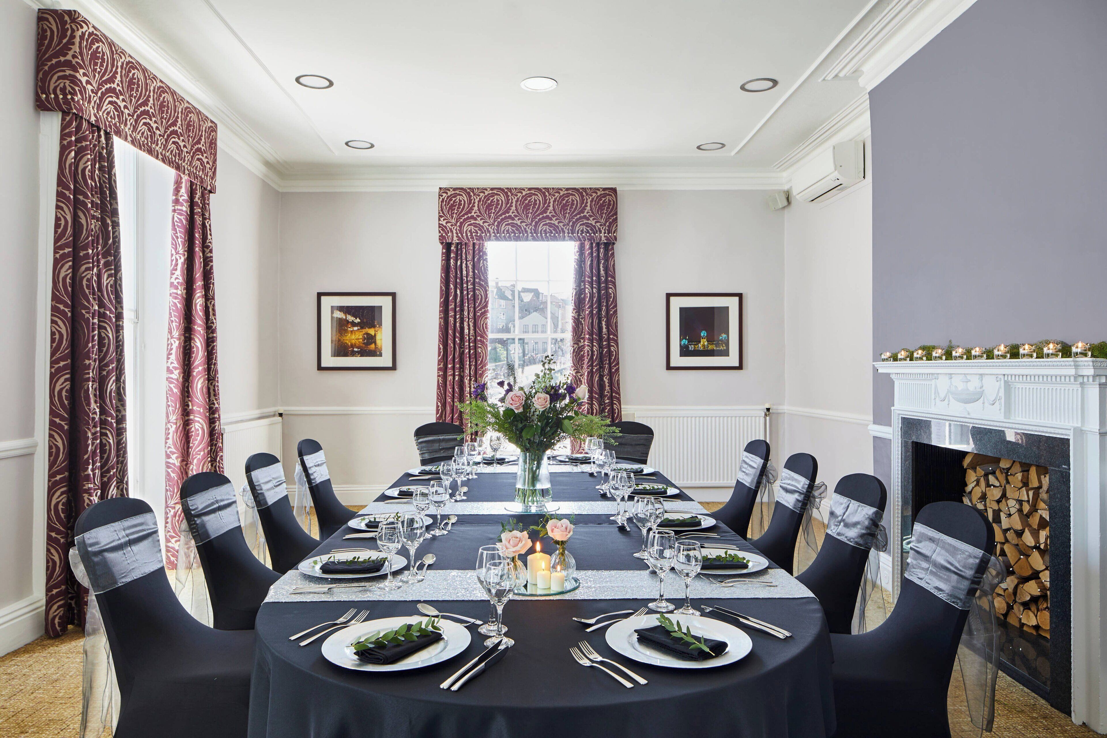 A formal dining room with a long table set for a meal, chairs with sashes, a vase with flowers, and a fireplace.