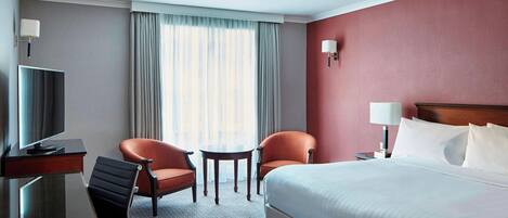 A hotel room with a bed, two armchairs, a small table, a desk with a TV, and a lamp.