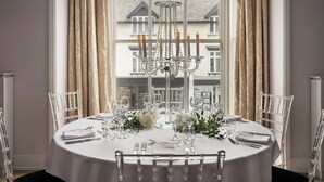 Mesa de comedor preparada para seis personas con sillas transparentes, una araña y vista a un edificio a través del ventanal.