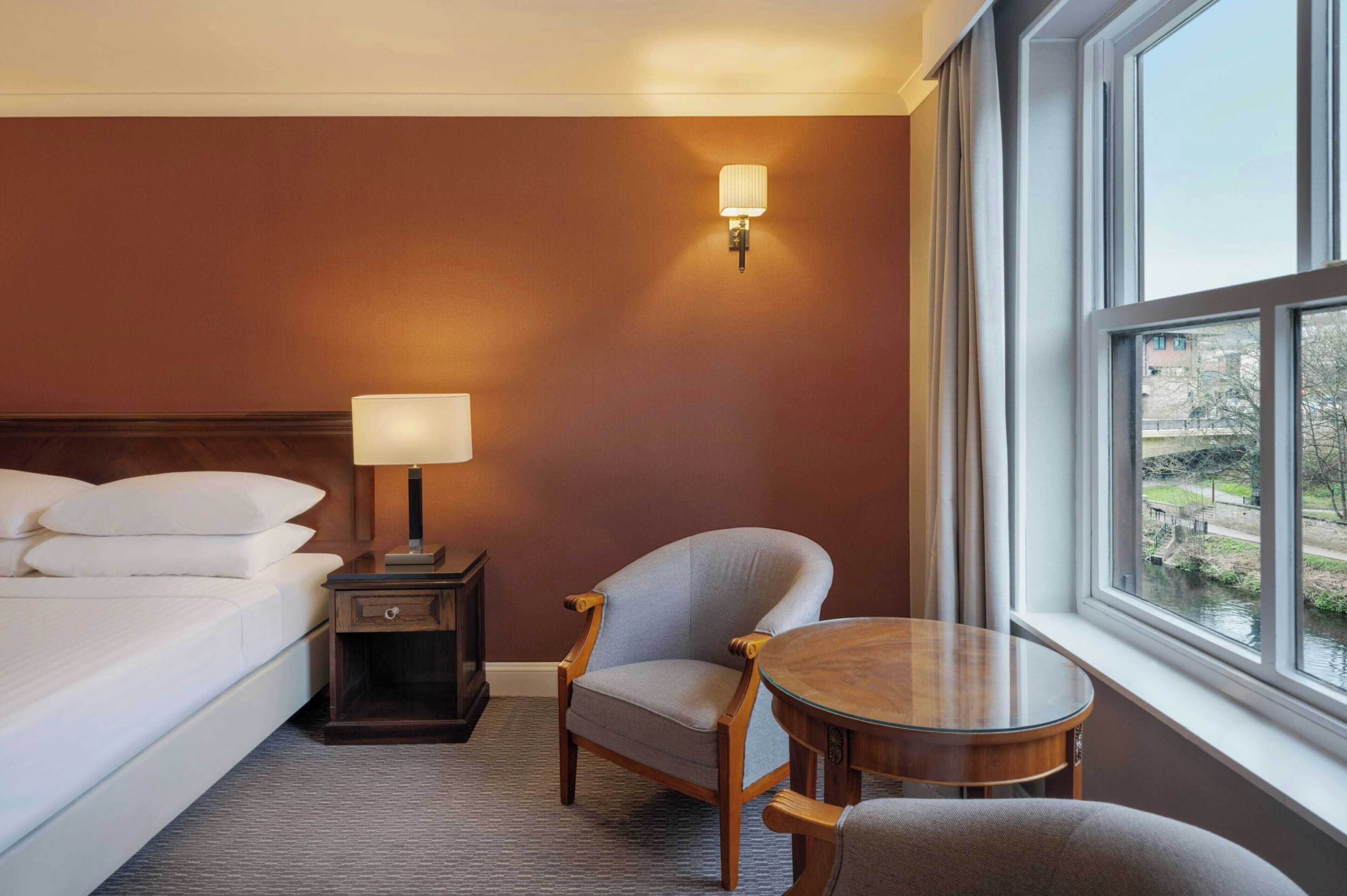 A hotel room with a bed, a chair, a small table, a nightstand, a lamp, and a window with a view of a river.